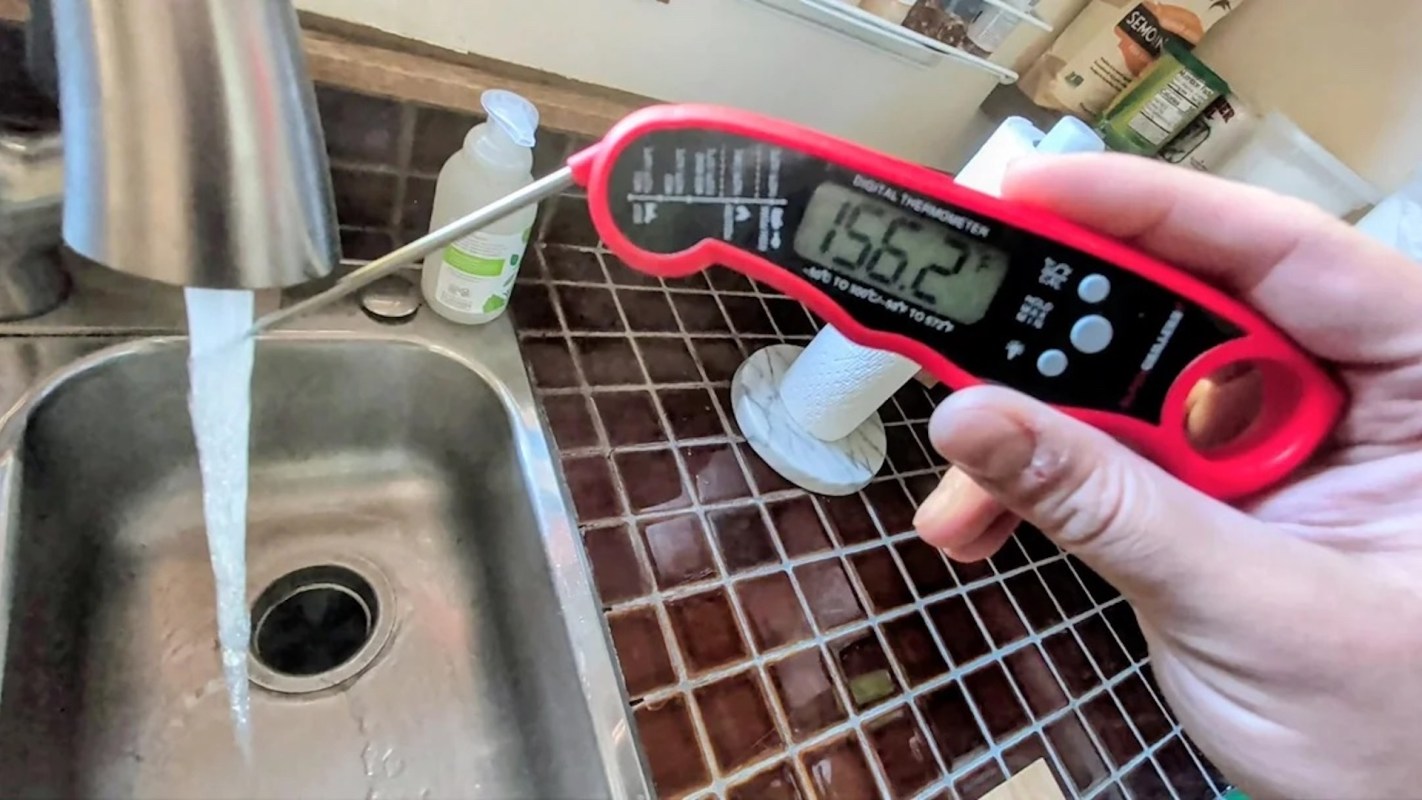 A hand holds a digital thermometer displaying a temperature reading of 156.2°F above a sink with running water.