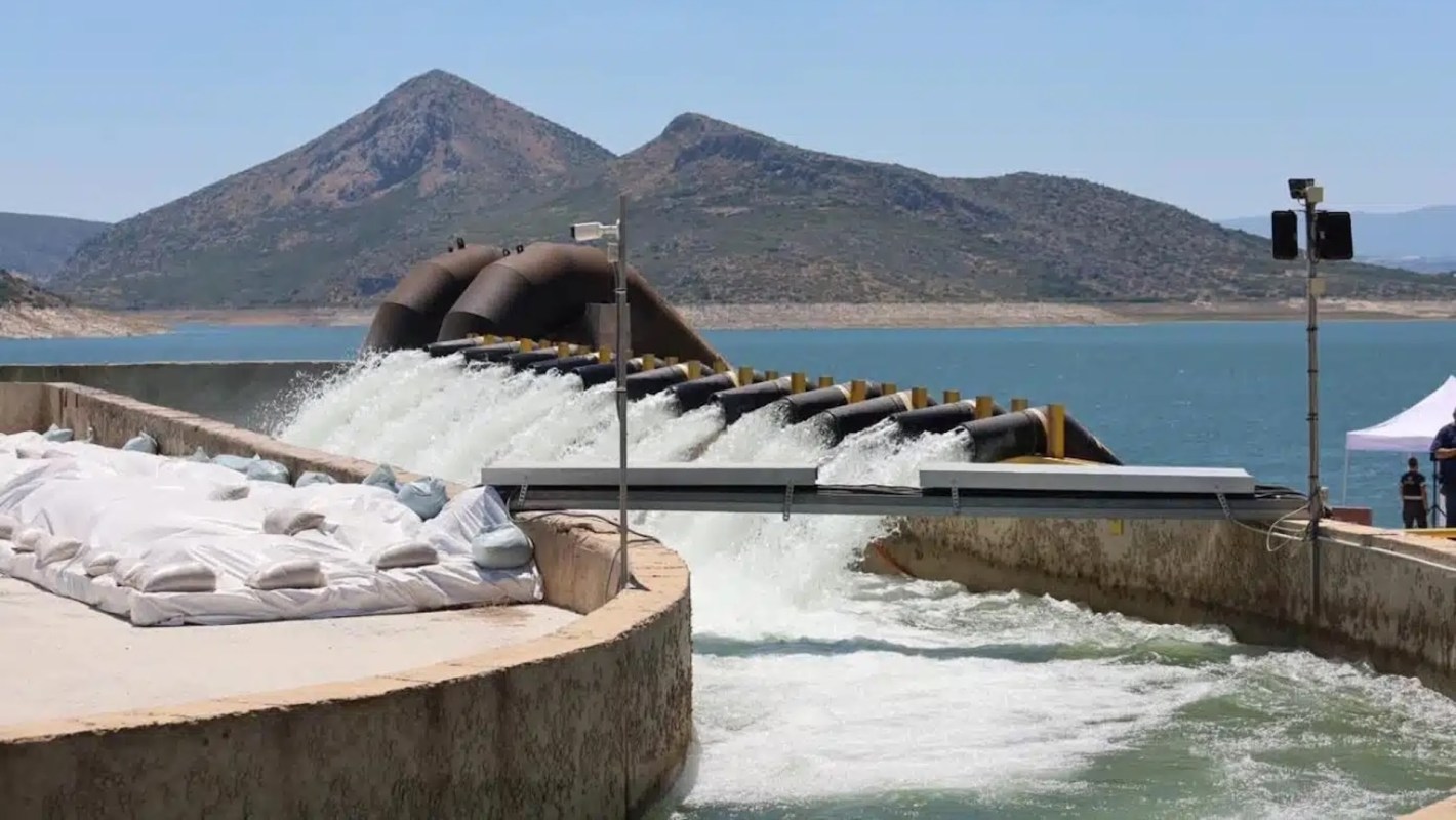 Water flows from a pipe into a reservoir of a treatment plant with mountains in the background.
