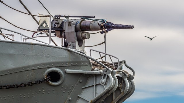 A ship's bow displays a mounted harpoon and ropes against a cloudy sky with a bird flying in the background.