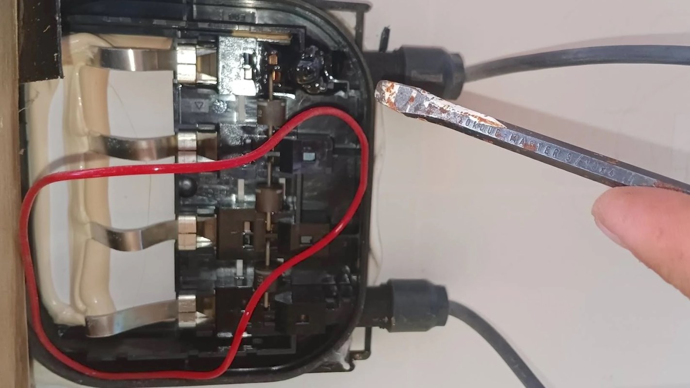 An electrical panel with wires and a rusted screwdriver pointing toward the connections inside.