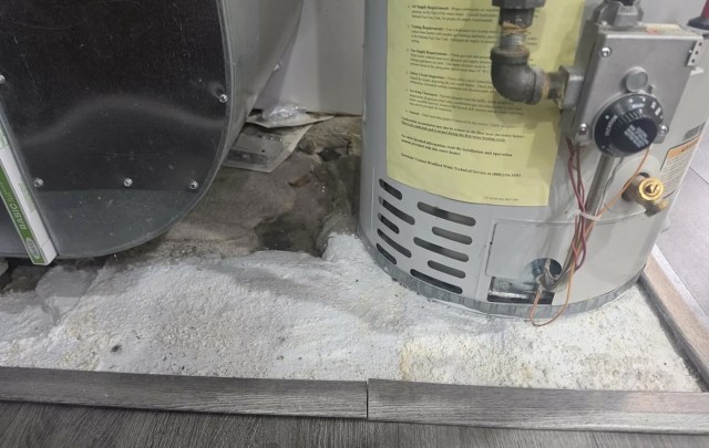 A water heater and metal vent near a patch of white material on a concrete floor.