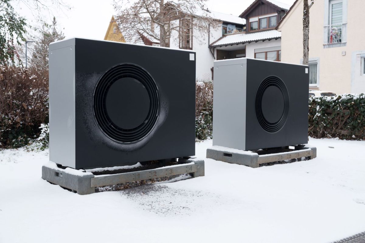Two heat pumps installed on concrete bases, covered in snow, surrounded by shrubbery and residential buildings.
