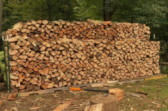 A large stack of firewood arranged neatly in a wooded area, with an axe resting on a log.