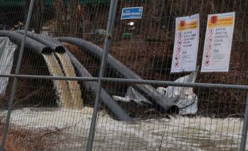 Two large pipes discharge brown water into a flooded area, surrounded by a chain-link fence and warning signs.