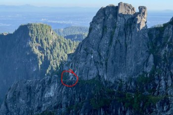 A rocky cliffside with dense trees and mountains in the background, with a red circle highlighting a stranded hiker.