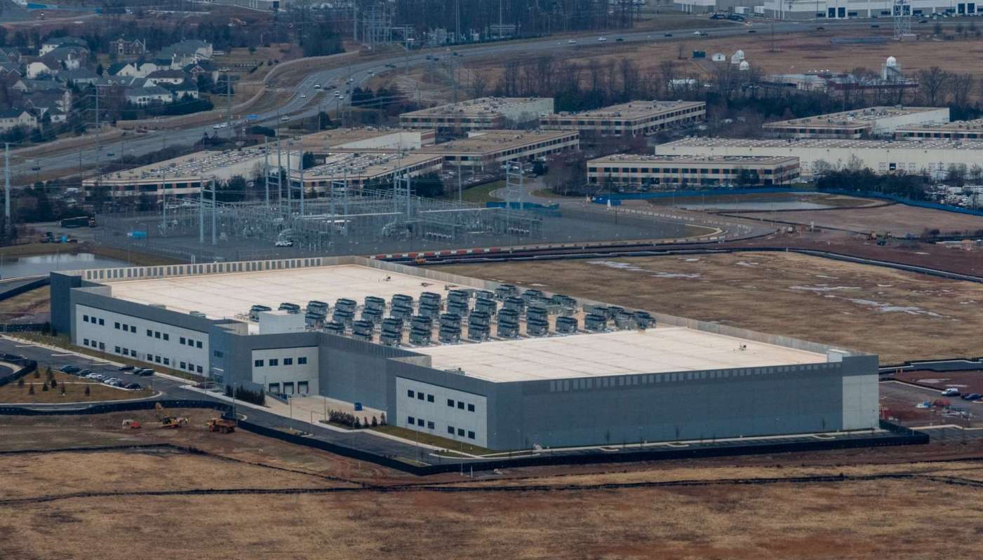 A large industrial building with rooftop equipment and nearby vehicles on a landscape with sparse vegetation.