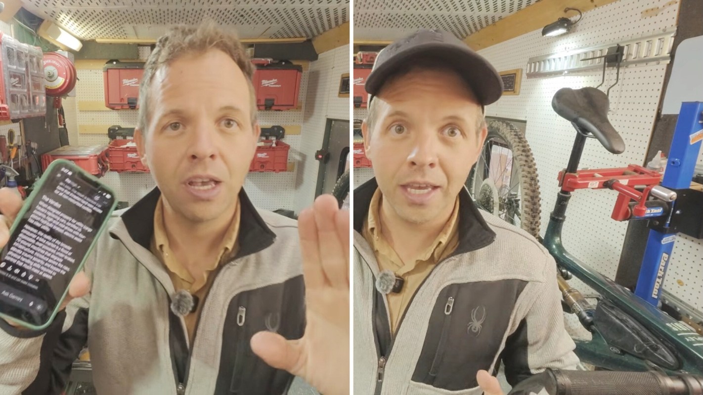 A man holds a smartphone and gestures in a workshop with tools and a bicycle in the background.