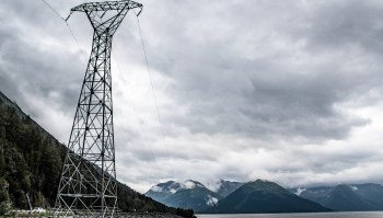 A power line tower stands near a lake, with mountains and cloudy skies in the background.