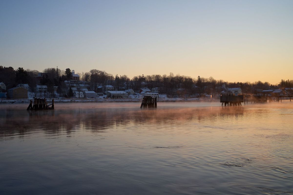 A calm river at Bucksport Maine at sunrise with mist rising and buildings along the shoreline.