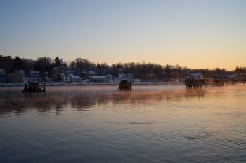 A calm river at Bucksport Maine at sunrise with mist rising and buildings along the shoreline.
