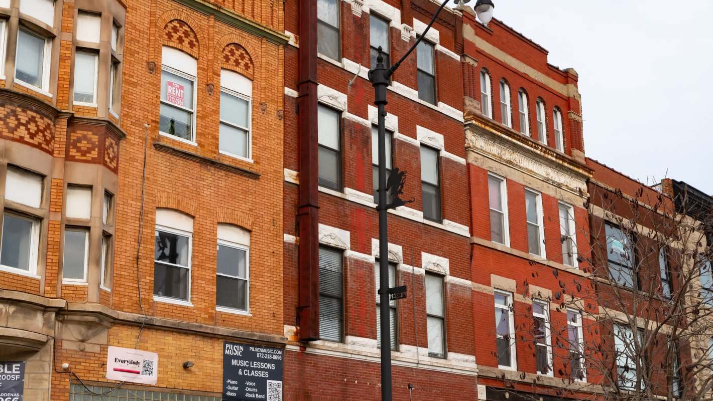 Brown and orange brick buildings with various windows and a "For Rent" sign. A streetlamp is visible in front.