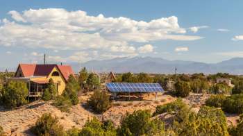 A house with a red roof and solar panels mounted on the ground, surrounded by trees and mountains under a partly cloudy sky.