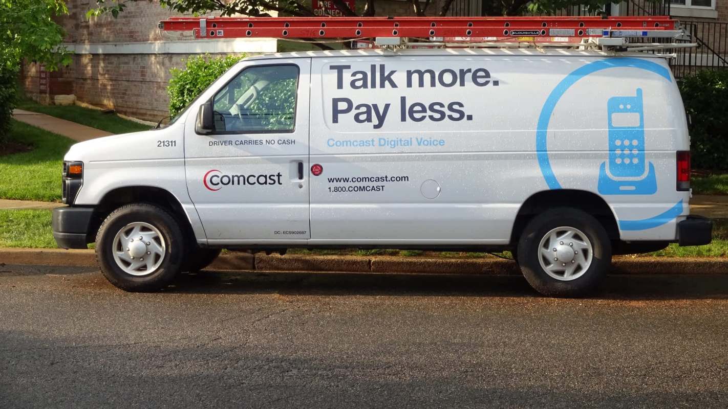 A Comcast service van parked on the street, featuring a ladder on the roof and promotional text on the side.