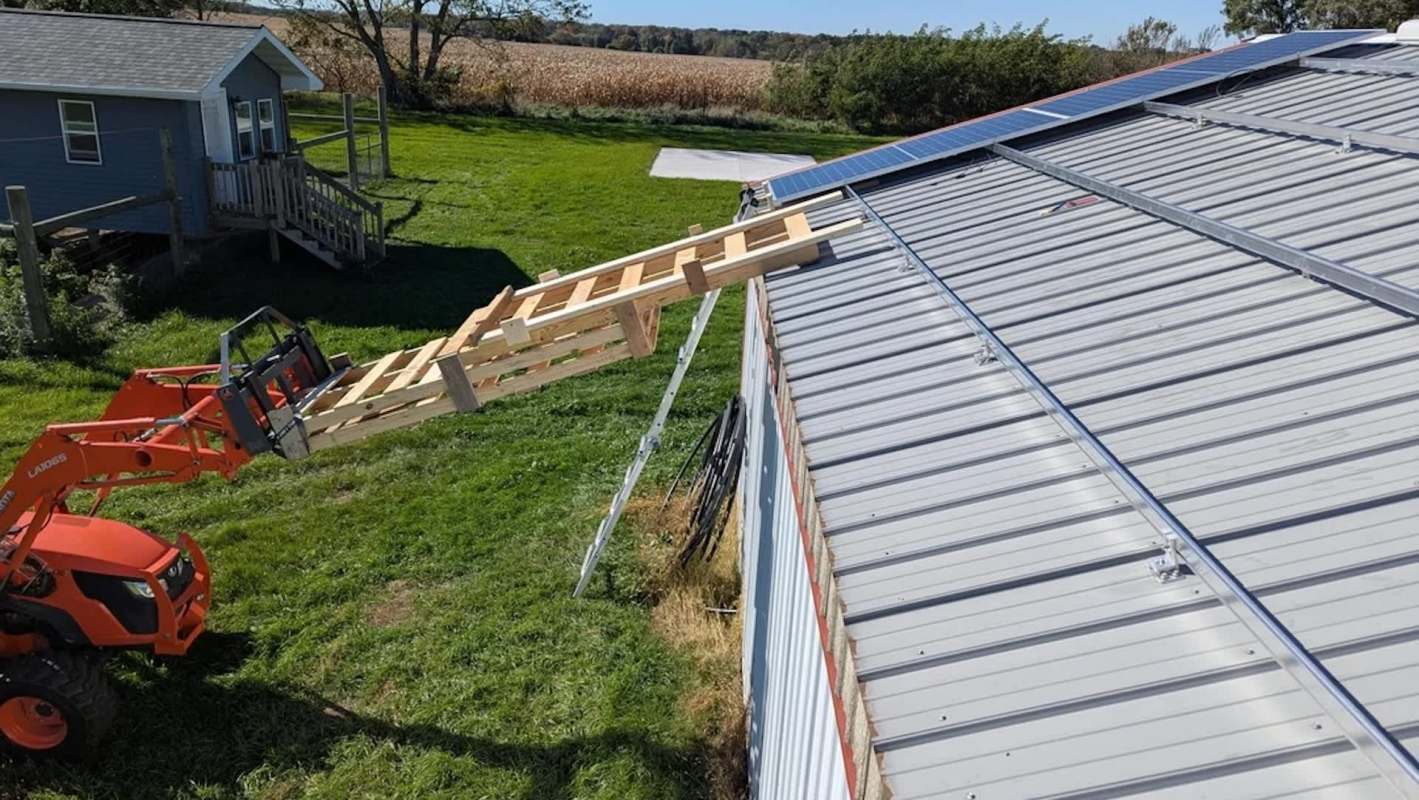 A loader with wooden planks is extended toward a metal roof with solar panels.
