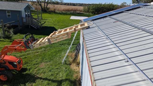 A loader with wooden planks is extended toward a metal roof with solar panels.