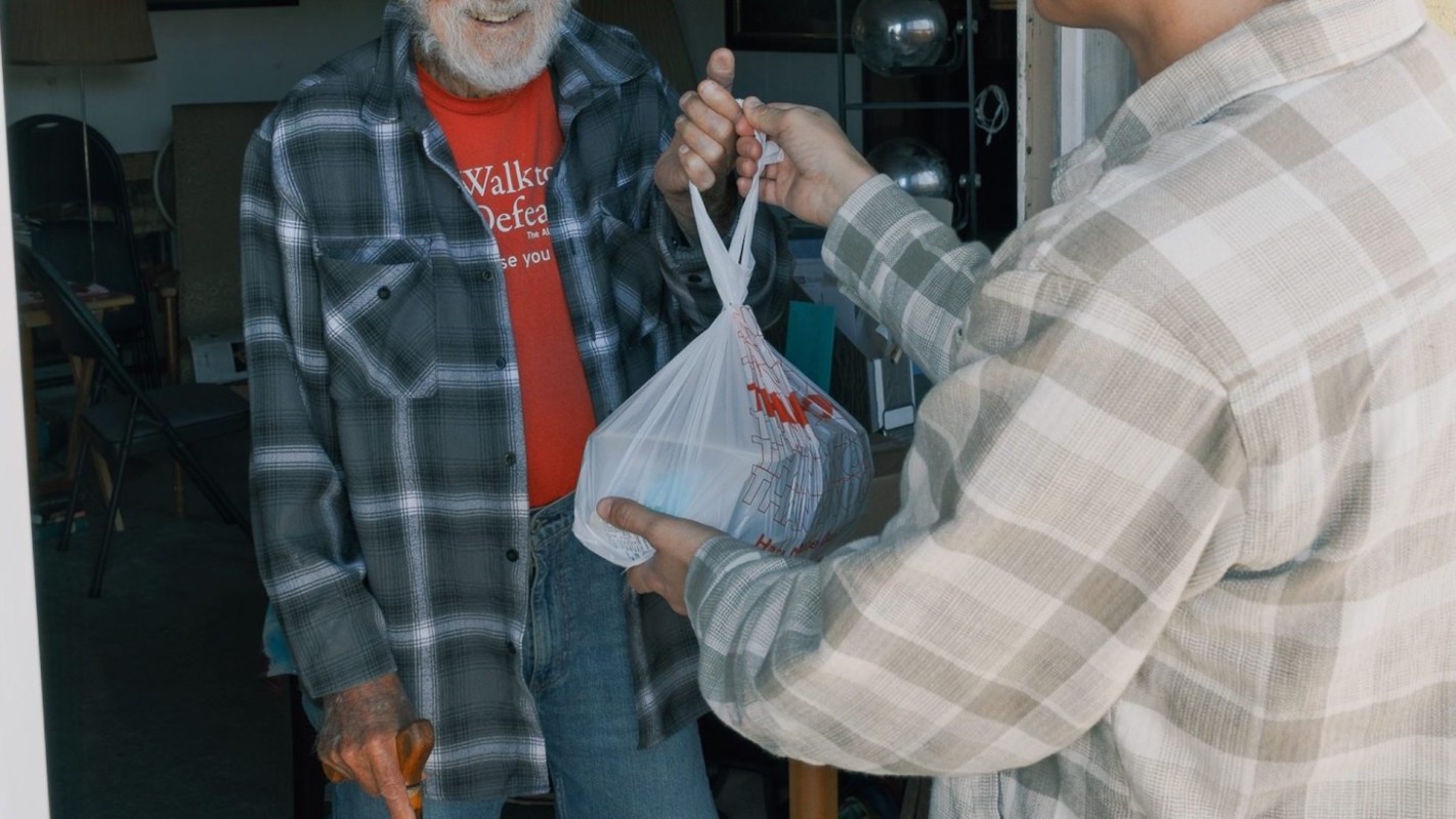 A person hands a bag to an elderly man standing at a door.