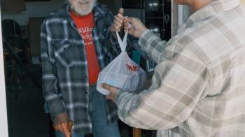 A person hands a bag to an elderly man standing at a door.