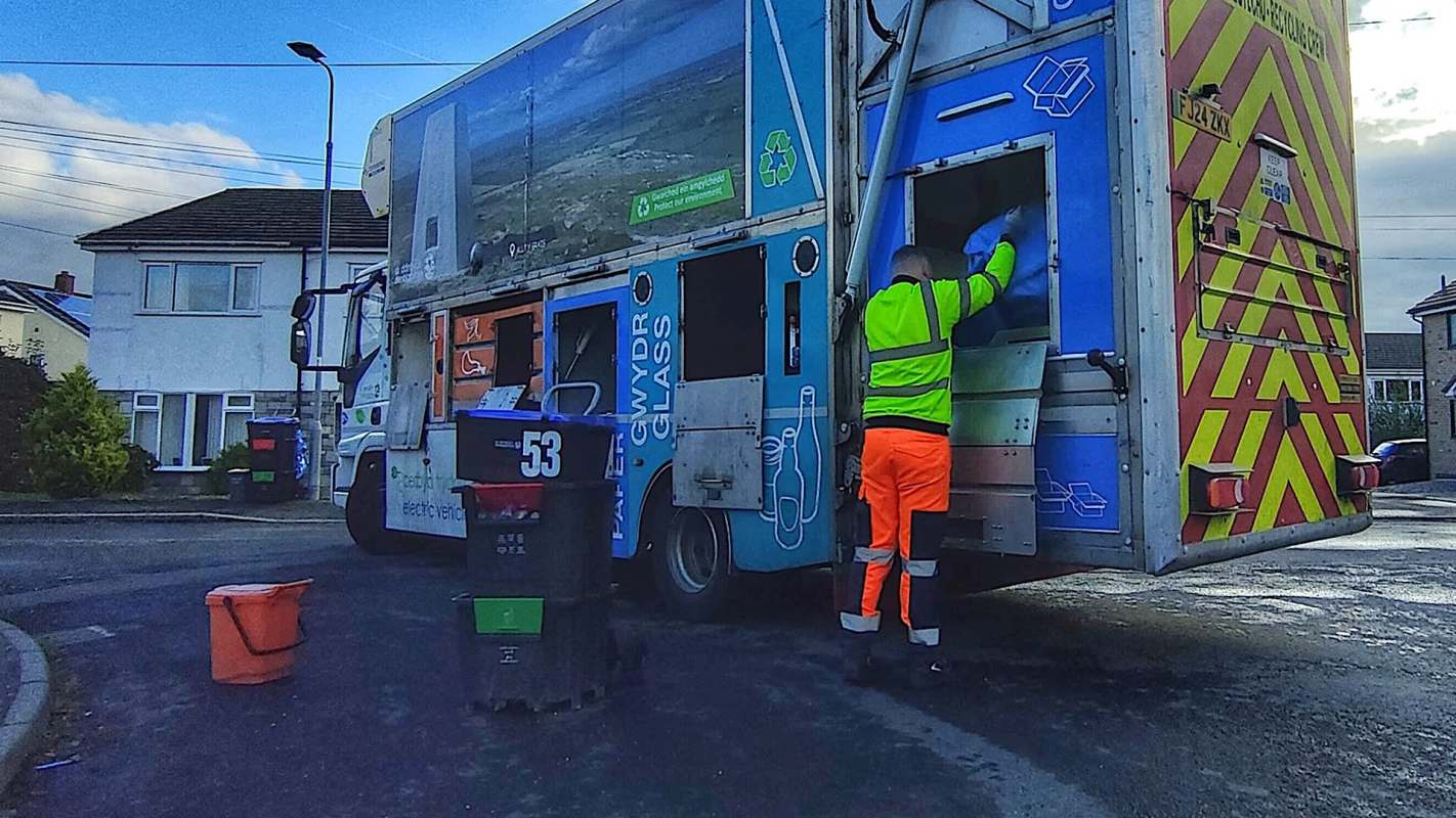 A person wearing a reflective vest empties a recycling truck at the curbside near residential homes.