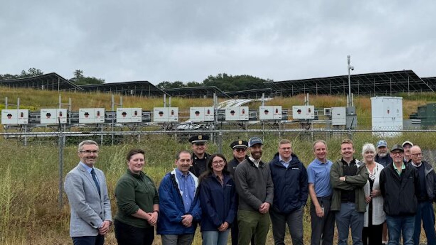 A group of people stands in front of a solar energy facility with electrical equipment in the background.