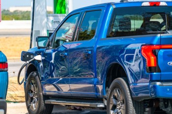 A blue electric Ford F-150 lightening truck is charging at a station with signage in the background.