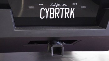 A black vehicle's rear with a custom license plate reading "CYBRTRK" and a tow hitch visible.