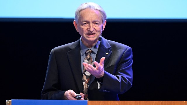 A man speaks at a podium, holding a remote control and gesturing with his hand.