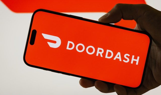 A hand holding a smartphone displaying the DoorDash logo on an orange background.