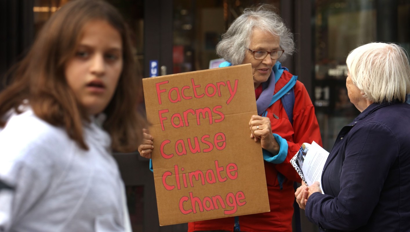 A woman holds a sign that reads "Factory Farms Cause Climate Change" while talking to another woman.