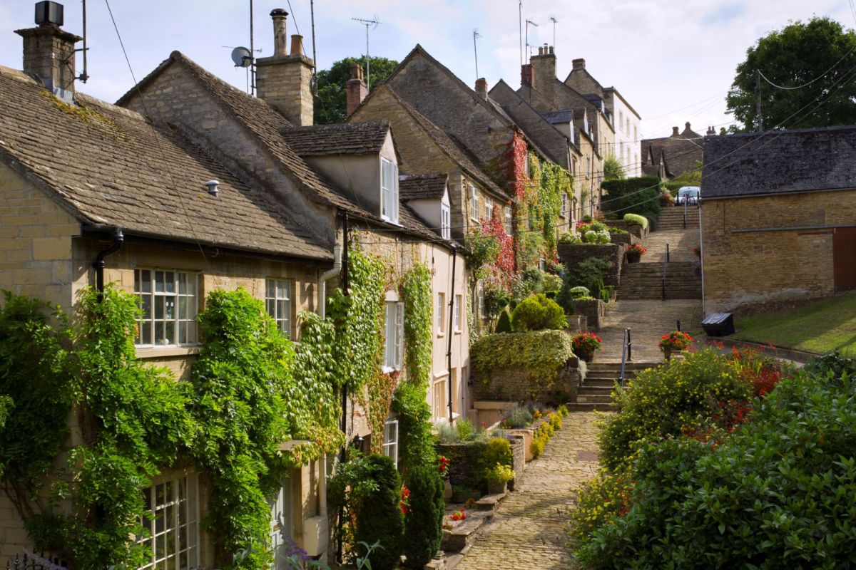A row of stone houses in Gloucestershire on a cobblestone path surrounded by greenery and flowers.