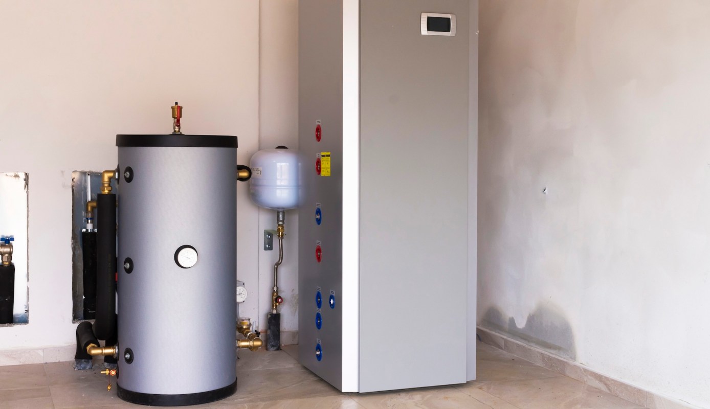 A gray water heater and a vertical tank stand against a wall in a utility space.