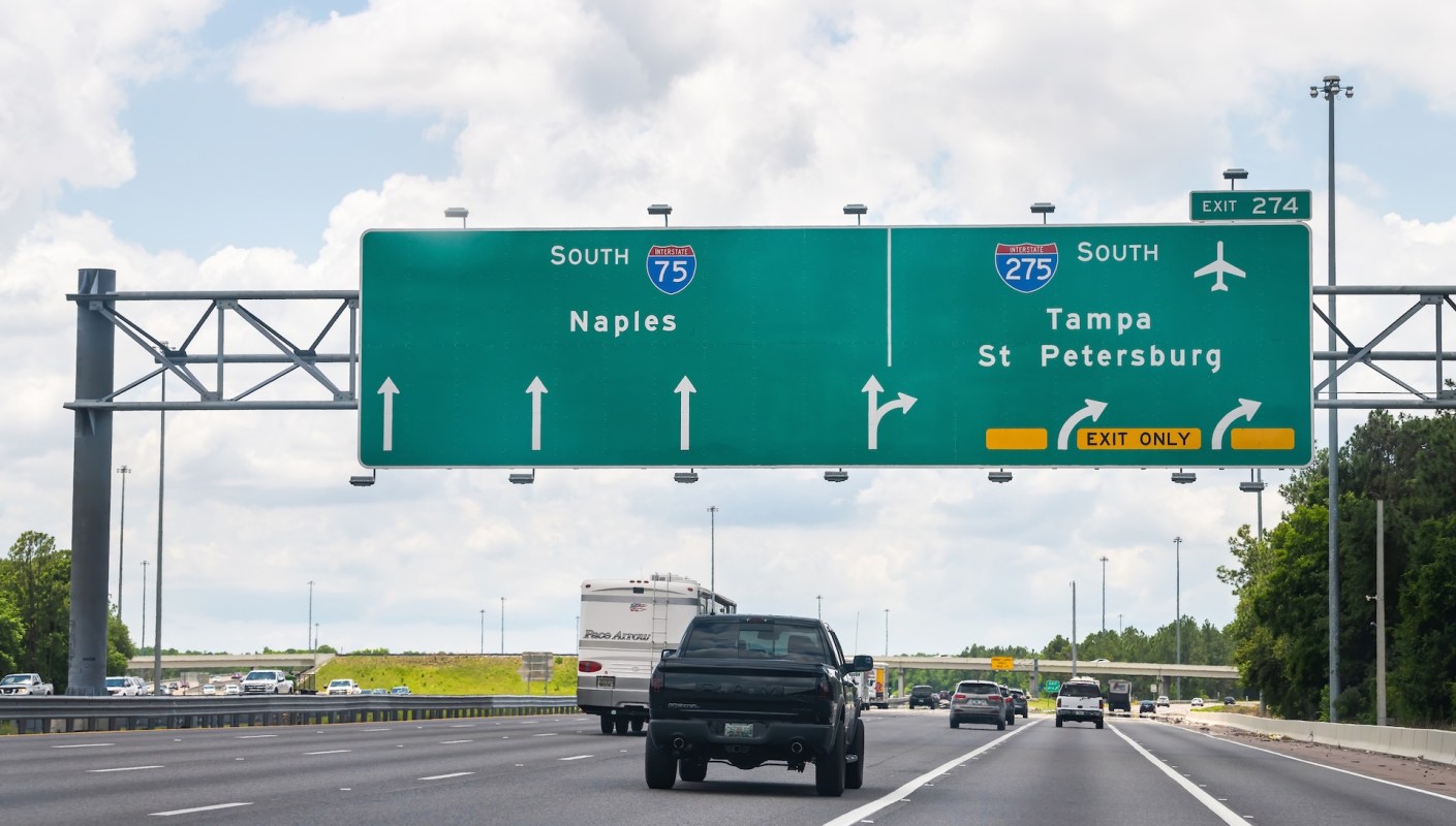 A highway sign indicating directions to Naples, Tampa, and St. Petersburg with vehicles on the road below.