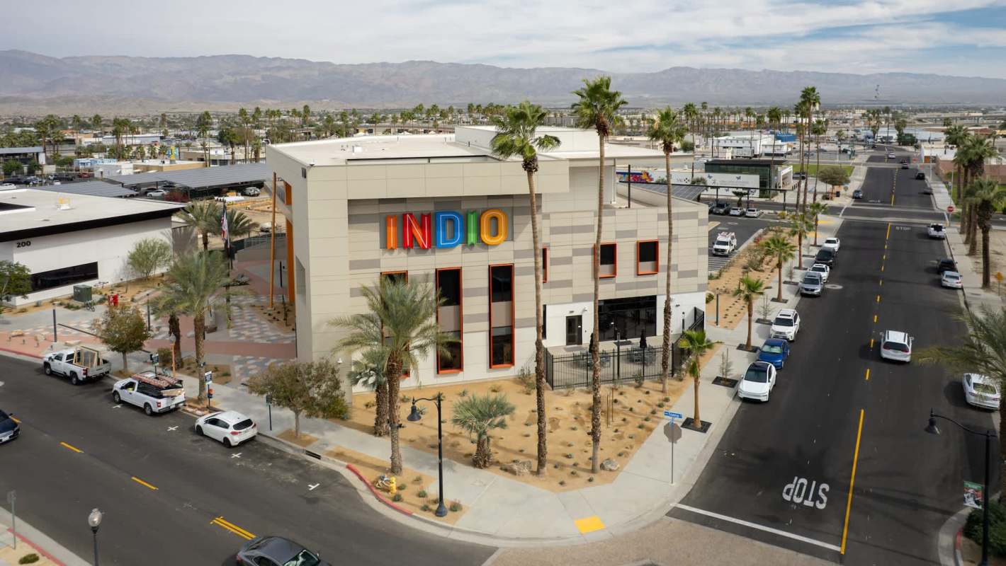 A modern building with colorful "INDIO" signage and palm trees, surrounded by a street and parked cars.