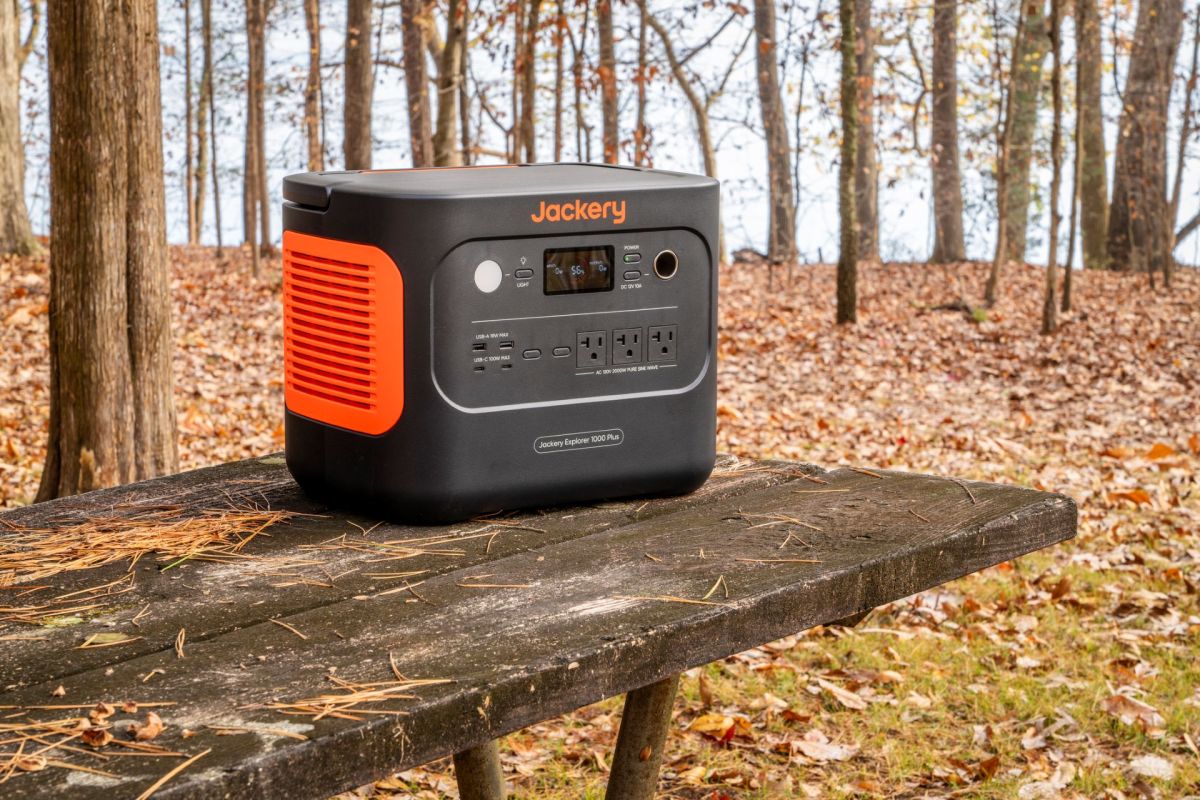 A portable Jackery power station on a wooden picnic table surrounded by trees and autumn leaves.