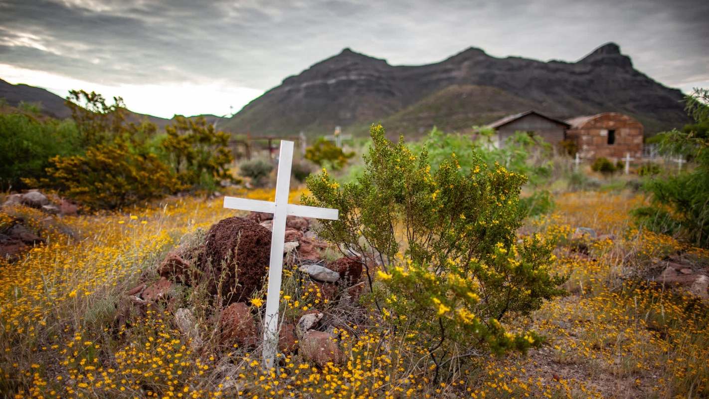 A white cross among yellow wildflowers with mountains and an abandoned building in the background.