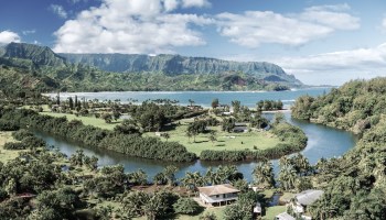 A mountainous landscape in Kauai with a river and coastal views, surrounded by lush greenery and residential areas.