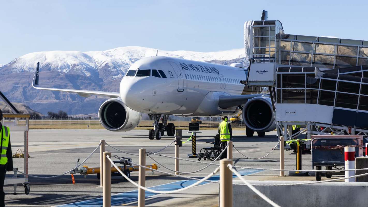 An Air New Zealand airplane is parked at the airport near a boarding bridge, with mountains in the background.