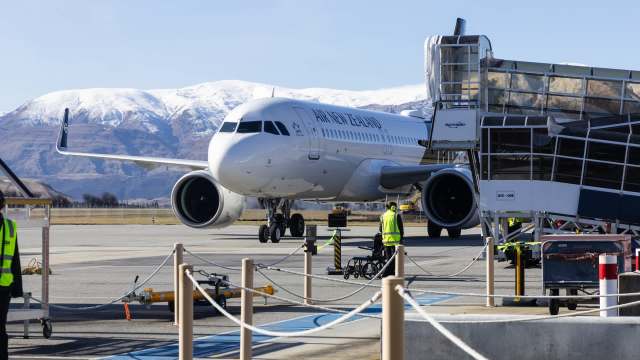 An Air New Zealand airplane is parked at the airport near a boarding bridge, with mountains in the background.