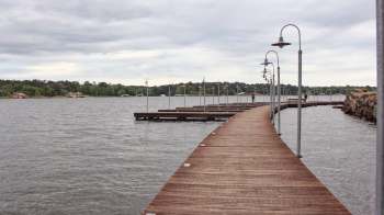 A wooden boardwalk lined with lamps extends over a lake, with trees and houses in the background.