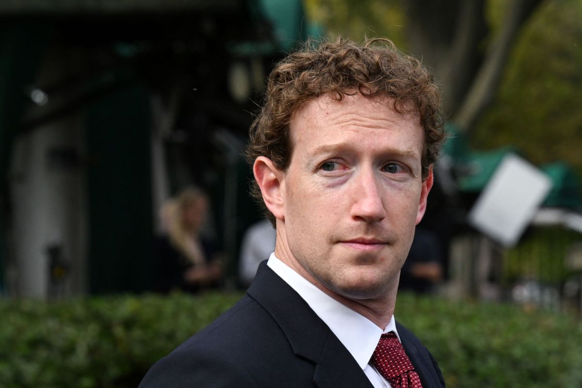 Mark Zuckerberg wears a suit standing outdoors near a hedge.