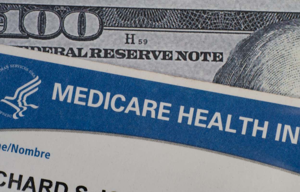 A Medicare health insurance card partially visible alongside a one-hundred-dollar bill.