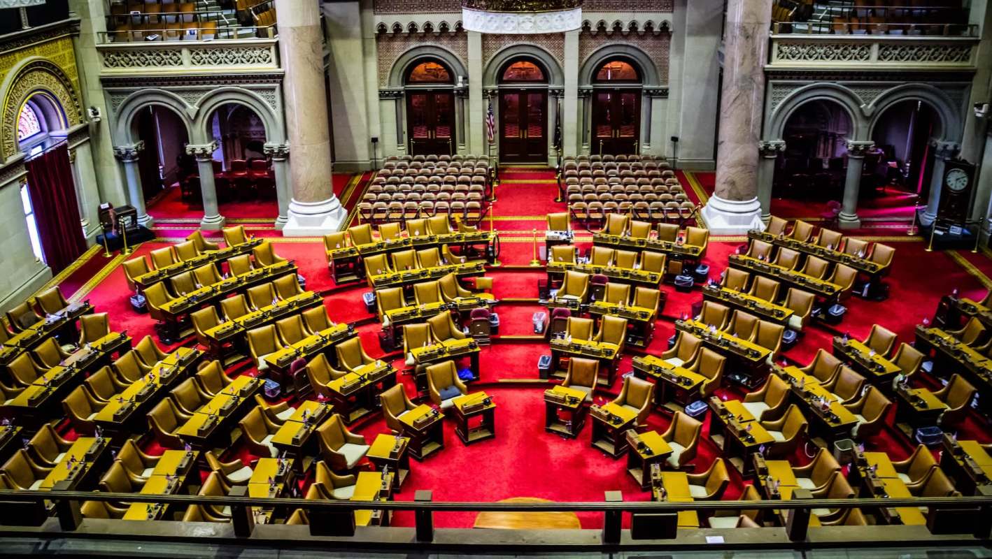 An empty legislative assembly chamber at the New York Capitol with rows of seats and a red carpet.
