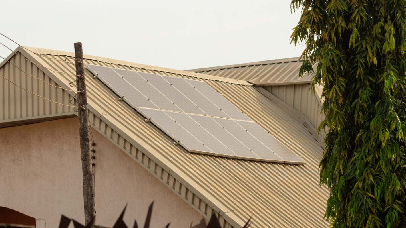 Solar panels installed on the roof of a building, with a tall tree nearby and electric poles in the background.