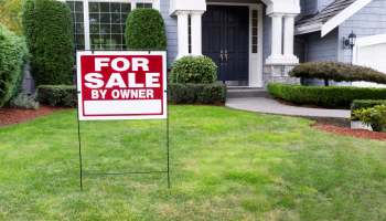 A "For Sale By Owner" sign stands in a grassy yard in front of a house.