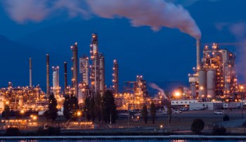 An industrial complex at night with illuminated structures and smoke rising from chimneys against a dark sky.