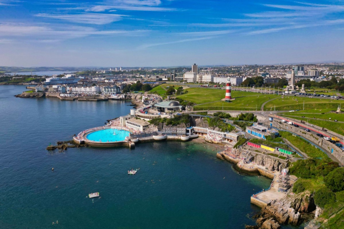 A coastal view featuring a swimming pool, several buildings, a lighthouse, and people enjoying the beach.