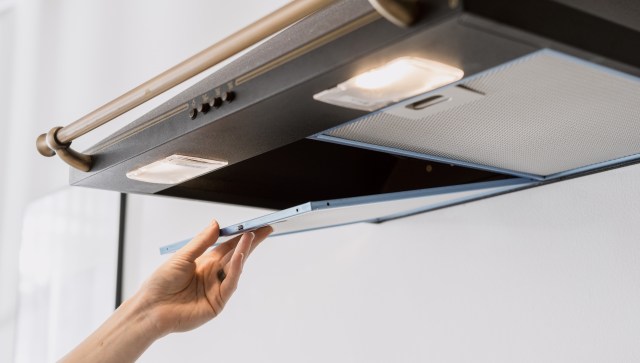 A hand is opening a kitchen range hood to access the filter.