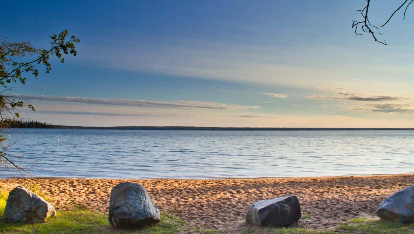 A sandy beach alongside a lake with rocks and trees under a clear blue sky.