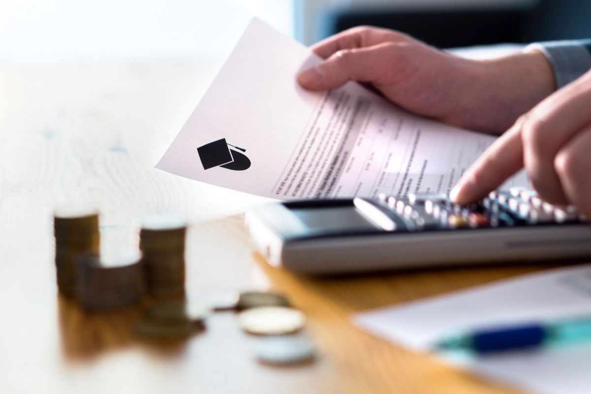 A person holds a paper with a graduation cap icon while using a calculator and coins are visible on the table.