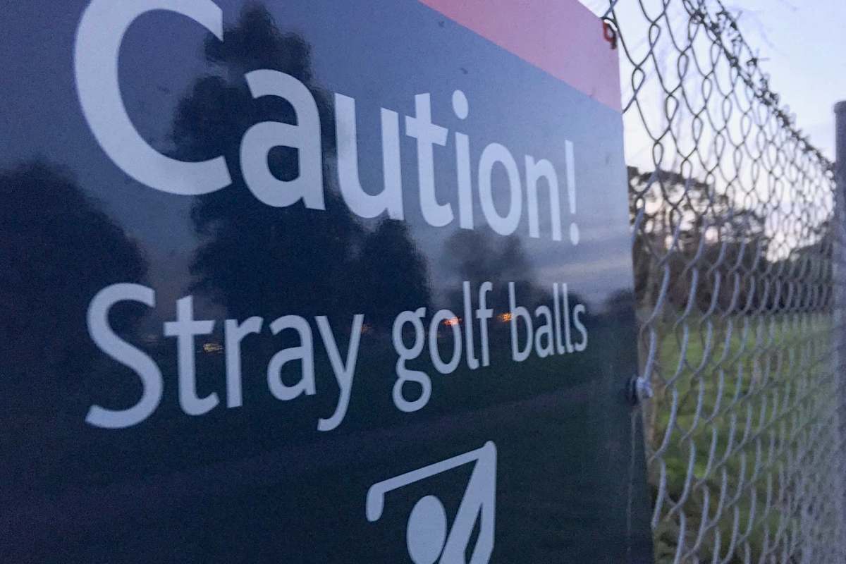 A warning sign stating "Caution! Stray golf balls" next to a chain-link fence.