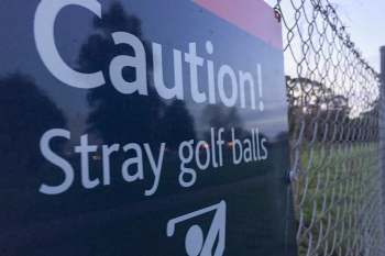 A warning sign stating "Caution! Stray golf balls" next to a chain-link fence.